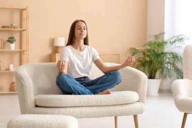 Genç ve güzel bir kadın oturma odasında kanepede meditasyon yapıyor.