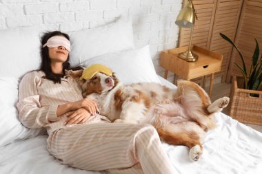 Avustralya çoban köpeği ve uyku maskeli genç bir kadın yatak odasında yatıyor.
