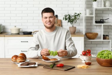Genç adam, mutfakta lezzetli bir burger için çöreğe marul koyuyor.