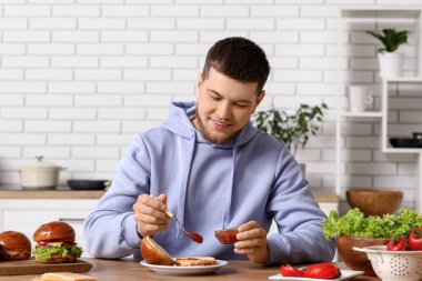 Genç adam mutfakta lezzetli bir burger için ekmeğe ketçap koyuyor.