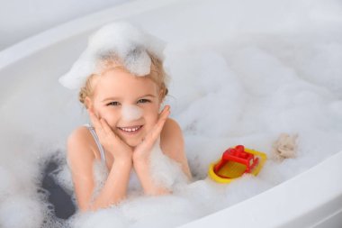 Şirin küçük kız banyo yaparken köpük ve oyuncaklarla banyo yapıyor.