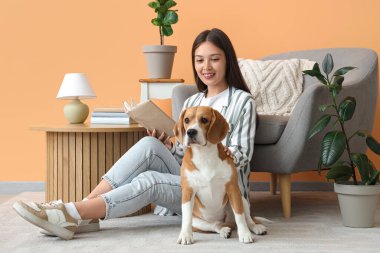 Oturma odasında yerde yatan sevimli Beagle köpeğiyle kitap okuyan genç bir kadın.