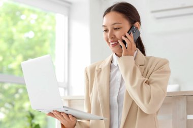 Modern dizüstü bilgisayarı olan genç bir bayan resepsiyonist, açık ofisteki resepsiyonda cep telefonuyla konuşuyor.