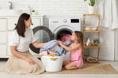 Çamaşır odasında çamaşır makinesinden yeni giysiler çıkaran genç bir kadın ve kızı var.