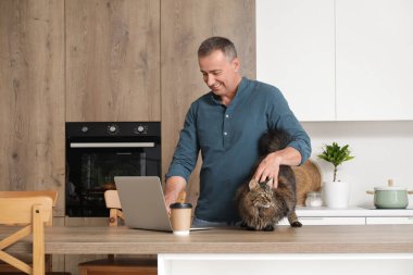 Evde dizüstü bilgisayarı ve kahve fincanı olan olgun bir adam masadaki sevimli kediyi okşuyor.