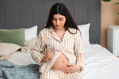 Evde pijamalı hamile bir kadın yatakta oturuyor.