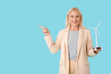 Rüzgâr türbini modeli olan olgun bir kadın mavi arka plandaki bir şeyi işaret ediyor.