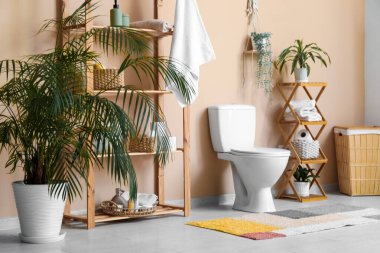 Beyaz klozetli modern tuvaletin içi, örülmüş sepet ve bej duvarın yanındaki raf ünitesi.