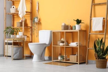 Beyaz klozet, sehpa ve turuncu duvarın yanında raf ünitesi olan şık bir tuvalet.