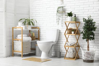 Beyaz tuğla duvarın yanında seramik klozet, ev bitkisi ve raf ünitesi olan modern tuvalet.