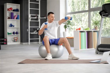 Sportif genç adam spor salonunda ağırlık kaldırma ve dizüstü bilgisayarla fitbol antrenmanı yapıyor.