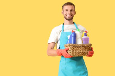 Sarı arka planda temizlik malzemeleriyle dolu sepeti tutan erkek hademe.