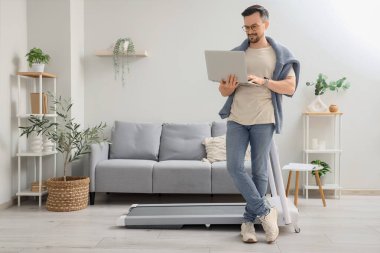 Evde koşu bandının yanında laptopla çalışan yakışıklı bir iş adamı.