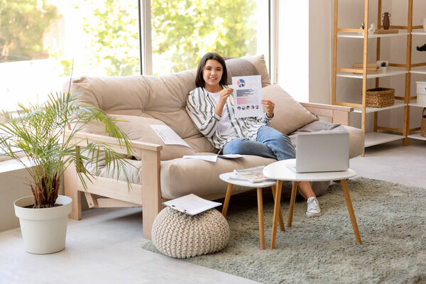 Молодая красивая деловая женщина с документами с графиками и сидя на диване в домашнем офисе