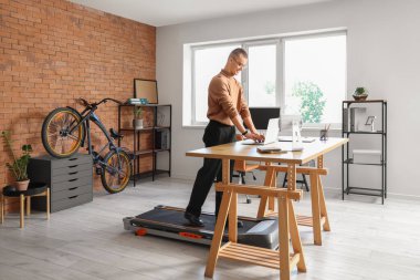 Genç işadamı ofisteki koşu bandında dizüstü bilgisayarla çalışıyor.