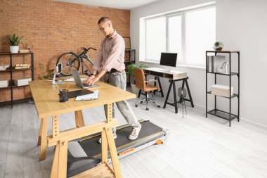 Genç işadamı ofisteki koşu bandında dizüstü bilgisayarla çalışıyor.