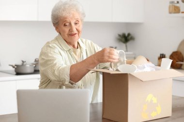 Mutfakta bir kutu çöp ve dizüstü bilgisayarı olan yaşlı bir kadın. Atık sıralama kavramı