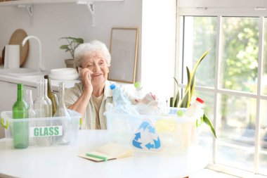 Mutfaktaki masada elinde çöp bidonlarıyla cep telefonuyla konuşan yaşlı bir kadın. Atık sıralama kavramı