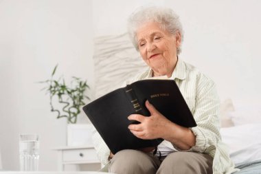 Yatakta yatan yaşlı kadın evde İncil okuyor.