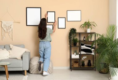 Oturma odasındaki bej duvarda boş bir çerçeve asılı genç bir kadın. Arka plan