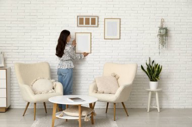 Evdeki beyaz tuğla duvara boş bir çerçeve asan genç kadın.