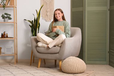 Evdeki katlanan perdenin yanındaki koltukta kitap okuyan genç bir kadın.