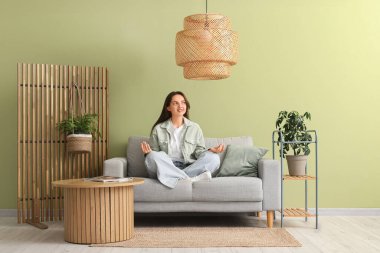 Evdeki gri kanepede meditasyon yapan genç ve güzel bir kadın.