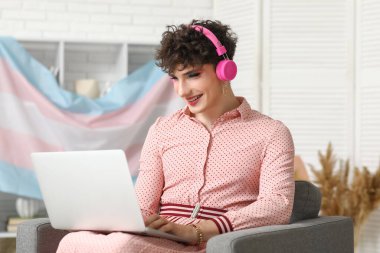 Kulaklıklı genç transseksüel kadın evdeki koltukta dizüstü bilgisayar kullanıyor.