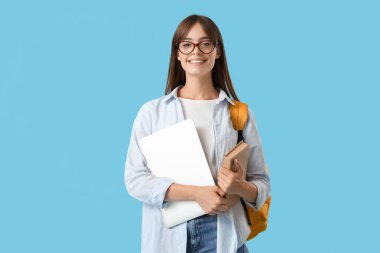 Mavi arka planda dizüstü bilgisayarı, kitabı ve çantası olan mutlu bir kız öğrenci.