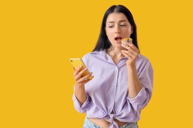 Cep telefonlu genç bir kadın sarı arka planda lezzetli kekler yiyor.