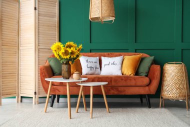 Yeşil duvarın yanındaki sehpanın üzerinde turuncu kanepesi ve vazo vazosu olan şık bir oturma odası.