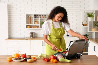 Güzel, genç, mutlu, Afro-Amerikan bir kadın mutfaktaki modern elektrikli ızgarada lezzetli sebzeler pişiriyor.