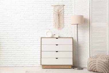 Çekmeceler, katlanır ekran, puf ve zemin lambası, beyaz tuğla duvarın yanında.