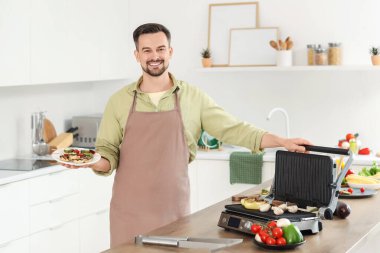 Mutfakta modern elektrikli ızgaranın yanında lezzetli sebzelerle dolu tabağı olan genç ve mutlu bir adam.
