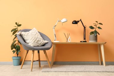 Turuncu duvarın yanında sazlık, saksı ve masa lambaları olan gri sandalye ve masa.