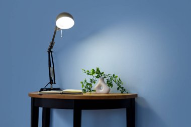 Üzerinde defter, çiçek ve mavi duvarın yanında parlayan masa lambası olan masa.