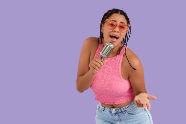 Güneş gözlüklü, mikrofonlu, leylak arka planda şarkı söyleyen genç bir Afro-Amerikalı kadın.