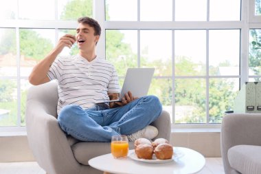 Dizüstü bilgisayarlı genç adam evdeki koltukta lezzetli çörekler yiyor.