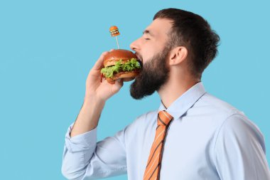 Genç işadamı mavi arka planda lezzetli bir burger yiyor.