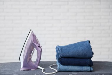 Beyaz tuğla duvarın yanında elektrikli ütü ve kot pantolon yığını.