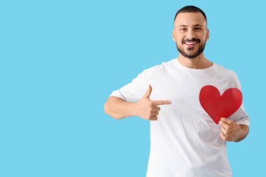 Yakışıklı genç adam mavi arka planda kağıt kalbe işaret ediyor.
