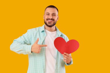 Yakışıklı genç adam sarı arka planda kağıt kalbe işaret ediyor.