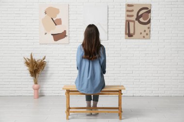 Sanat galerisinde beyaz tuğlalı duvardaki resimlere bakan genç bir kadın.