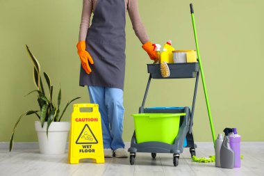 Kadın hademe, tramvay dolusu temizlik malzemesi ve yeşil duvarın yanında uyarı levhası taşıyor.