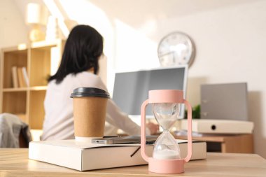 Ofiste çalışan iş kadınına karşı masasında kahve fincanı, dosya ve cep telefonu olan kum saati, yakın plan.