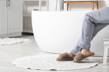 Evdeki küvetin yanında, halının üzerinde yumuşak terlikler içinde kadın bacakları, yakın plan.