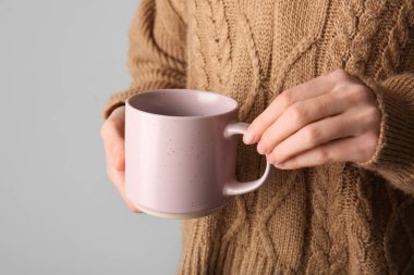 Gri arka planda seramik kupası olan şık örülü süveter giyen kadın, yakın plan.