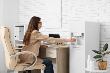 Güzel genç iş kadını modern su soğutucusunu açık ofiste kullanıyor.