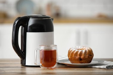 Modern elektrikli çaydanlık, bir bardak sıcak çay ve mutfaktaki ahşap masada tatlı bir pasta.