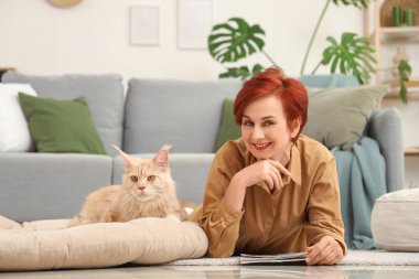 Evde yerde yatan sevimli Maine Rakun kedisi olan olgun kızıl saçlı kadın.